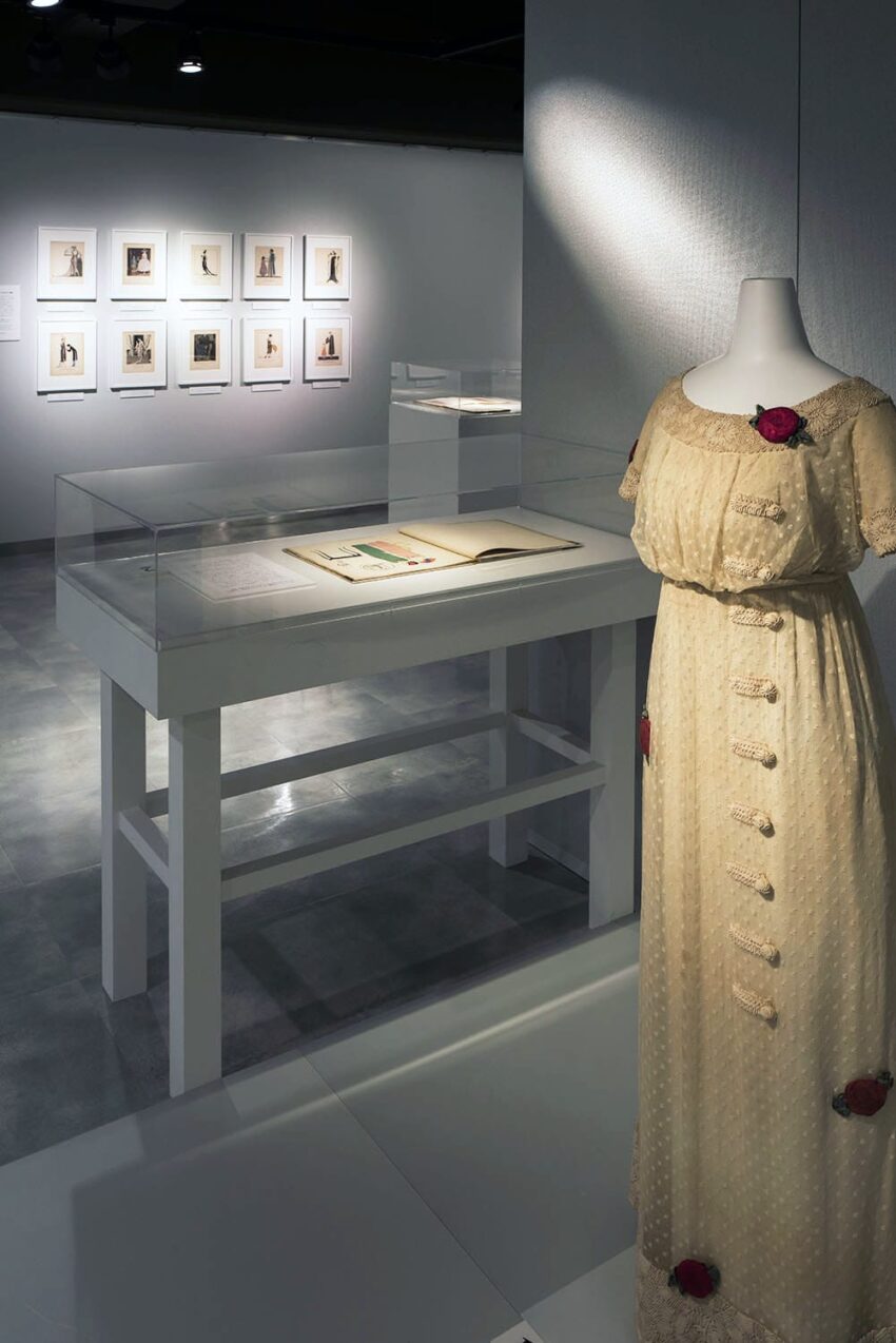 Museum display with a vintage dress on a mannequin, open book in a glass case, and framed photos on a wall.