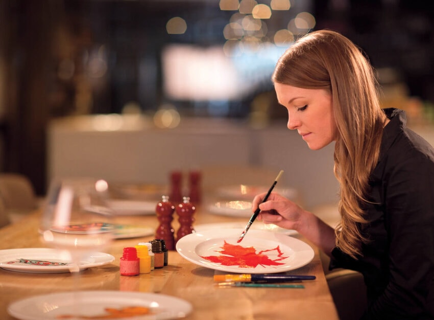 Woman painting a red maple leaf on a white plate at a wooden table with paints and brushes nearby.