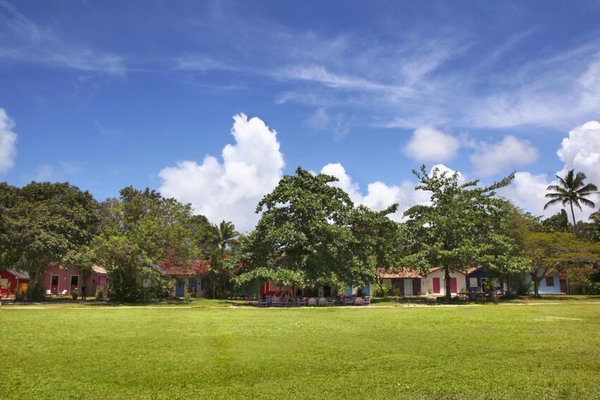 Colorful houses behind a green field under a vivid blue sky with fluffy clouds and lush greenery.