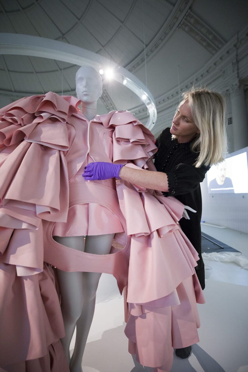 Designer adjusting a mannequin wearing an elaborate pink ruffled gown in a well-lit exhibition space.