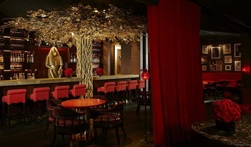 Luxurious bar interior with red seating, a golden statue, elegant lighting, and floral decor, creating a sophisticated ambiance.