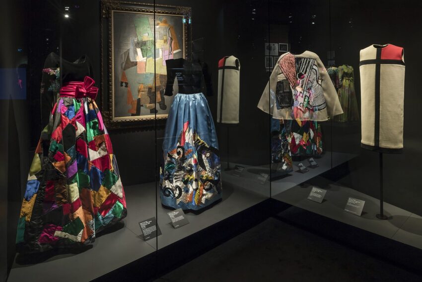 Colorful fashion exhibit featuring patchwork dresses, a painting, and garments displayed in a dimly lit museum setting.