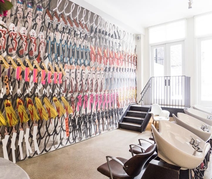 Hair salon interior with colorful art mural on wall behind several washing stations and chairs.