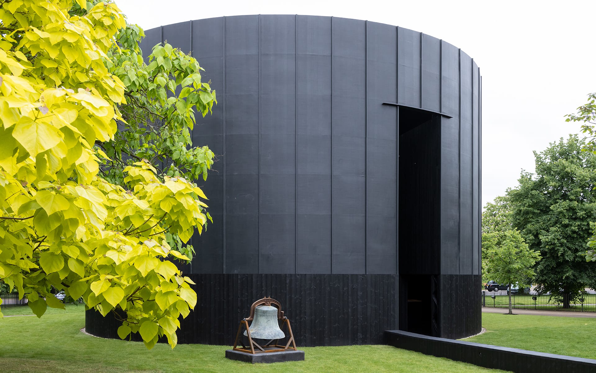 Round black building with curved walls and open doorway, surrounded by lush green trees and a bronze bell sculpture in front.
