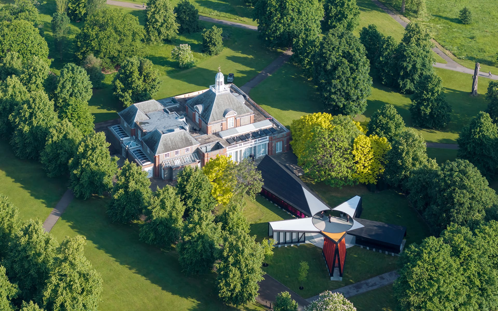 Aerial view of a historic building surrounded by lush trees in a park with modern architectural structure nearby.