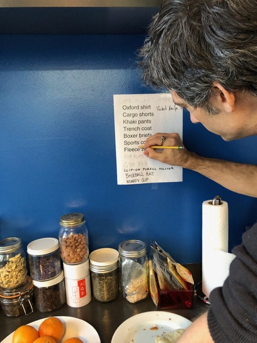 Person writing on a list pinned to blue wall above kitchen counter with assorted jars and oranges.