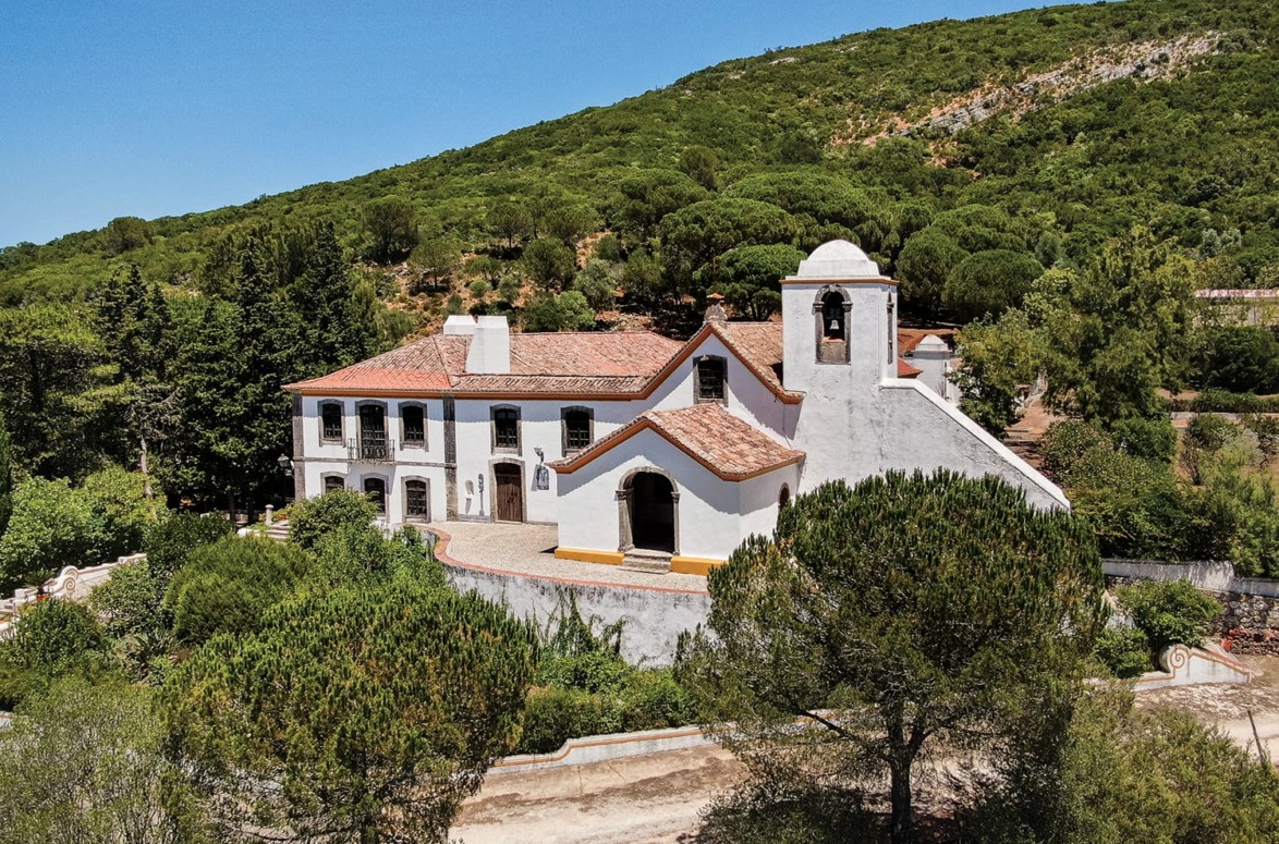 White villa with red roof surrounded by lush green trees and hills on a sunny day.