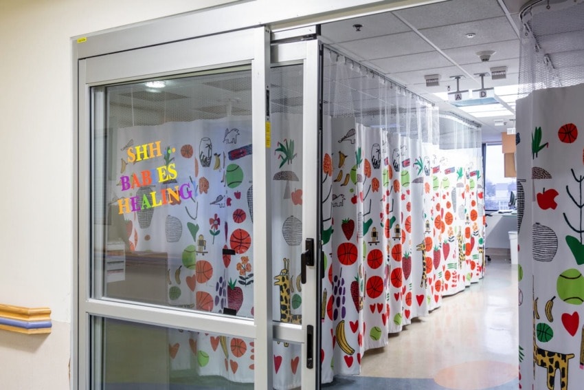 Hospital room with a sliding glass door labeled "Shh...Babies Healing" and colorful patterned privacy curtains inside.