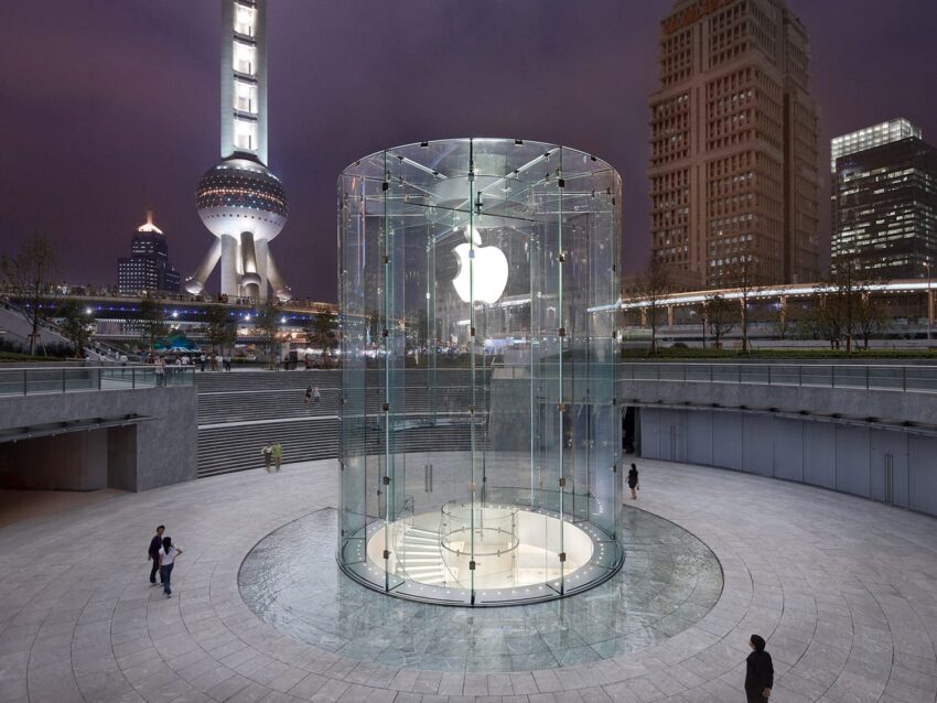 Glass cylinder entrance to an Apple Store surrounded by a plaza, with a view of city skyscrapers at night.