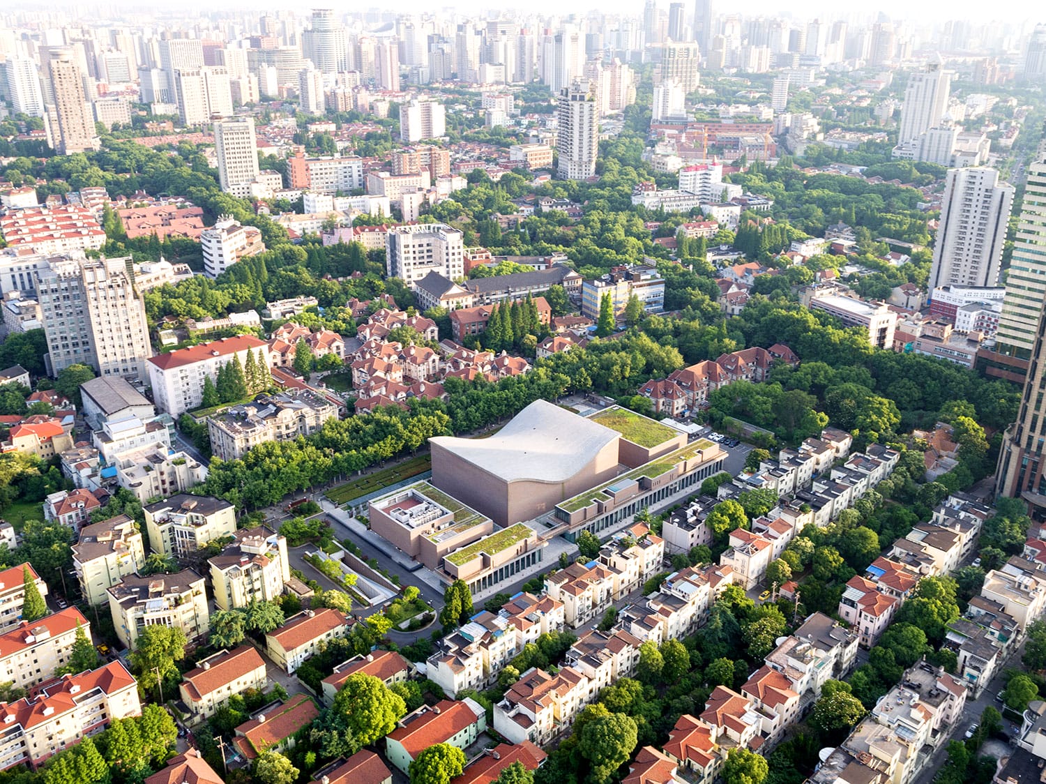 Aerial view of a modern building surrounded by green trees in an urban residential area with city skyline in the background.