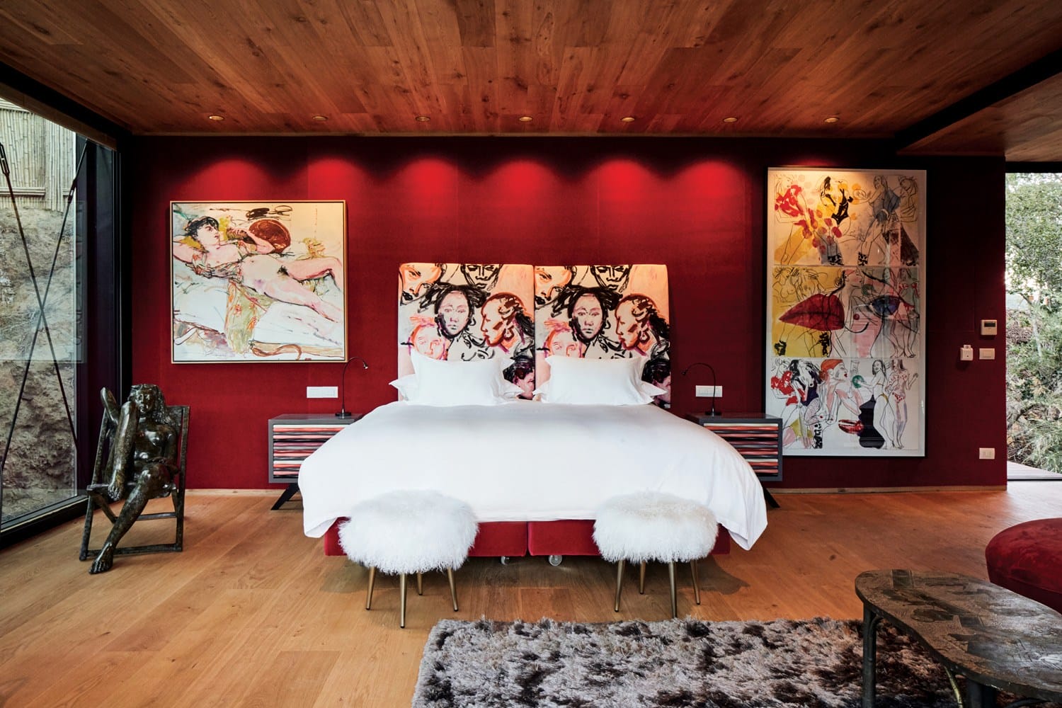 Modern bedroom with bold red walls, wooden ceiling, artistic paintings, white bed, fluffy stools, and a large window.