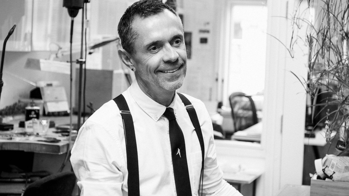 Smiling man in an office wearing a white shirt with black suspenders and a tie, sitting at a desk.