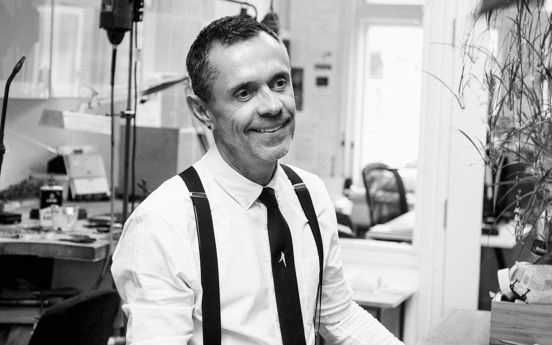 Smiling man in an office wearing a white shirt with black suspenders and a tie, sitting at a desk.