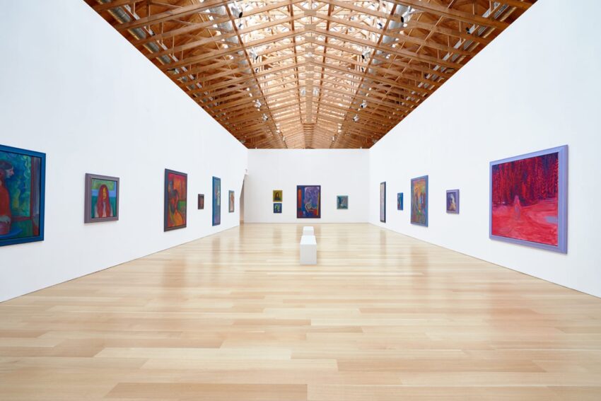 Modern art gallery with wooden ceiling, featuring various colorful paintings on white walls and a wooden floor.