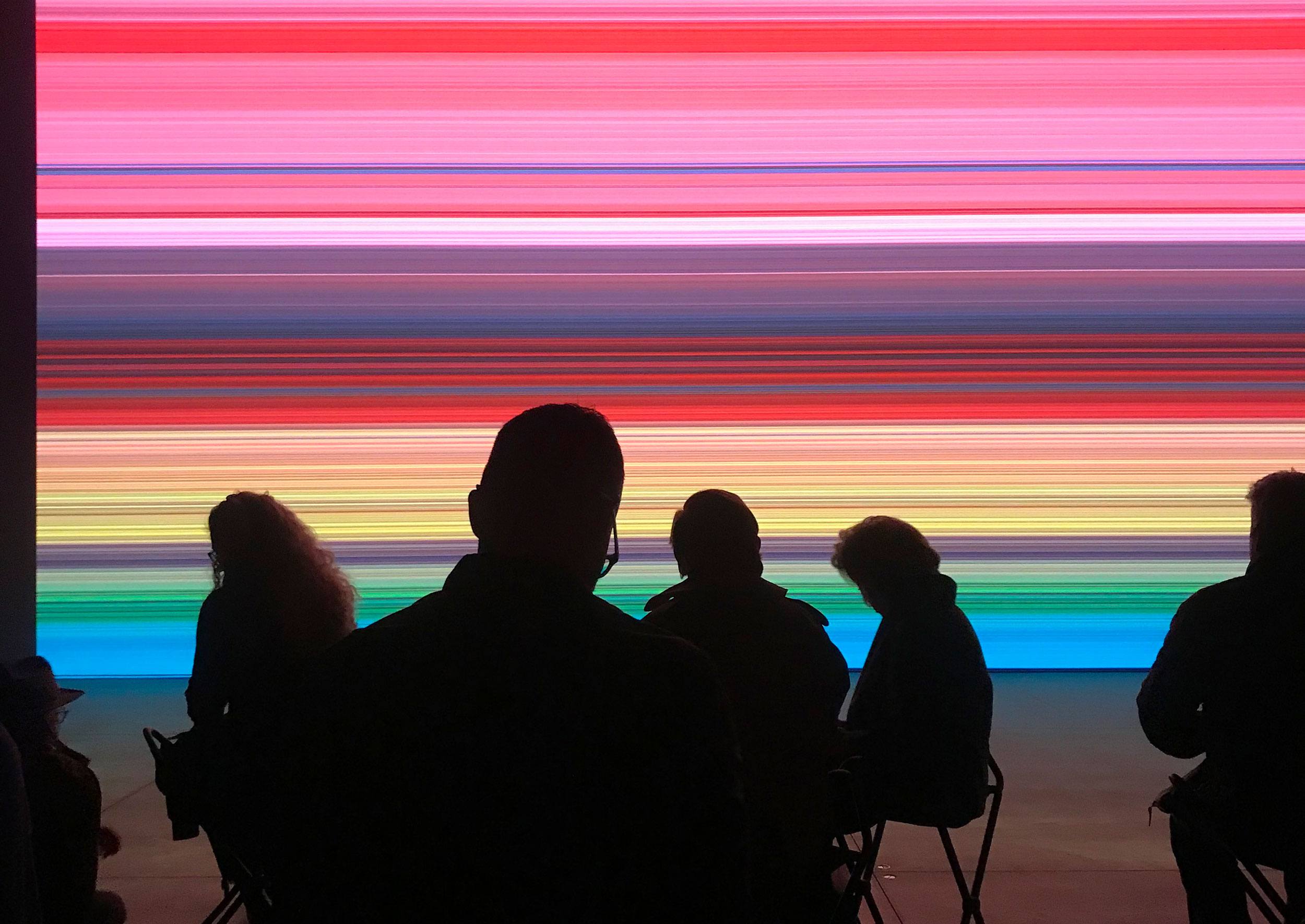 Silhouettes of people sitting in front of a vibrant, colorful digital art display with horizontal stripes.