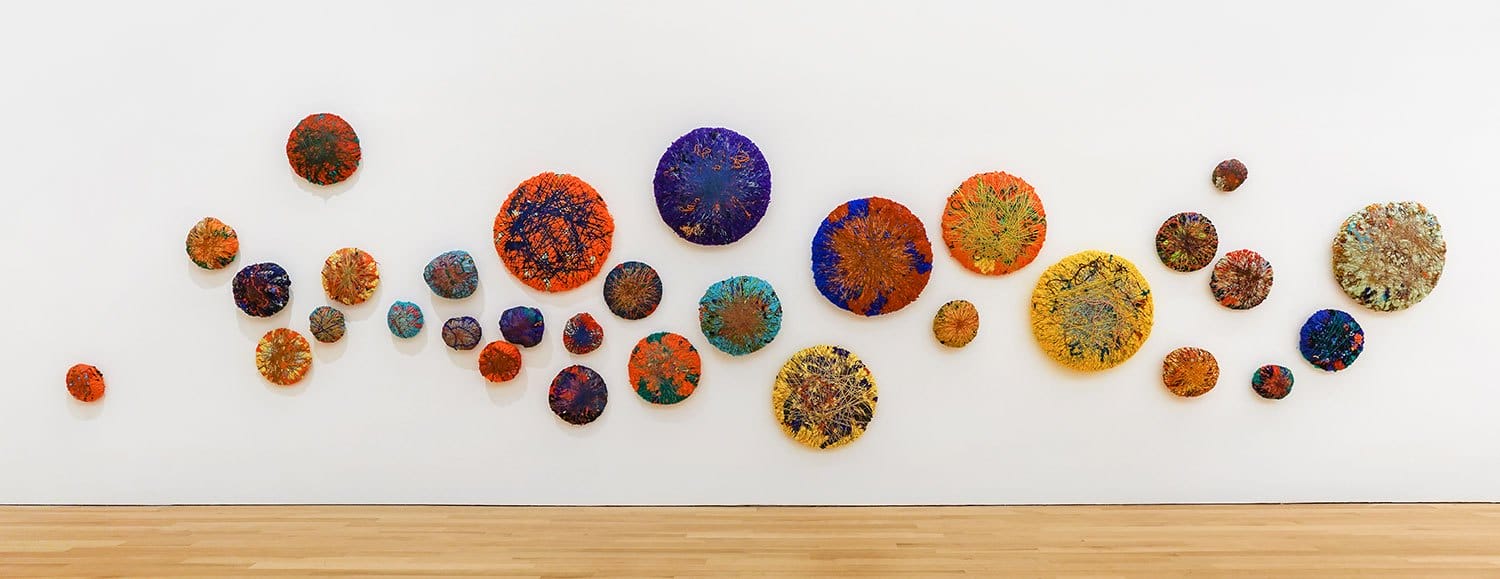 Colorful circular abstract art pieces displayed on a white gallery wall above a wooden floor.