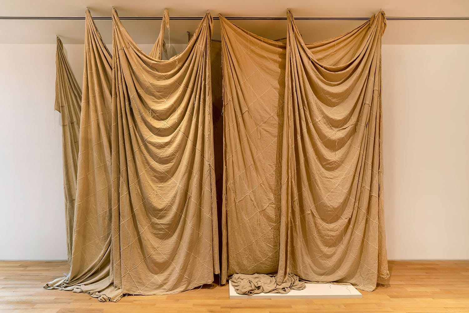 Large draped fabric installation in an art gallery setting, covering wall space from ceiling to floor on wooden floorboards.