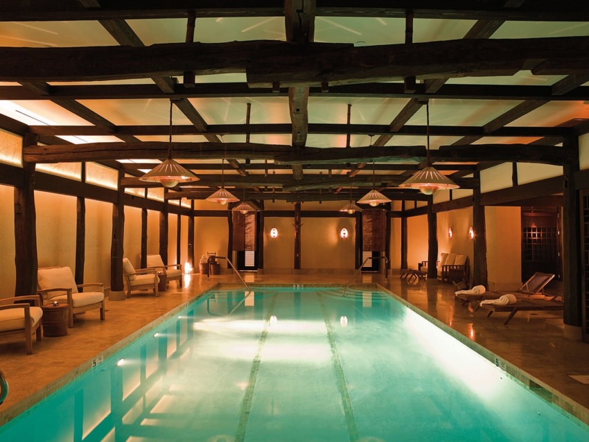 Indoor swimming pool with warm lighting, wooden beams, and lounge chairs along the sides, creating a relaxing atmosphere.