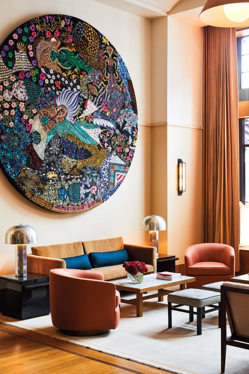 Modern living room with large colorful tapestry, brown sofa, two orange chairs, and tall windows with curtains.