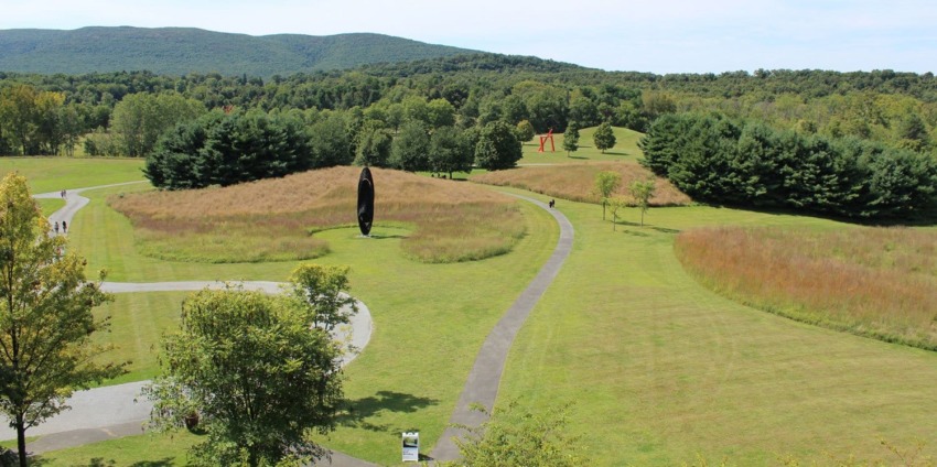 Outdoor sculpture park with winding paths, trees, grassy fields, and distant hills under a blue sky.