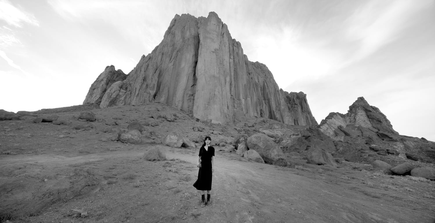 A person in a black outfit stands on a rocky terrain before a massive rock formation under a cloudy sky, black and white photo.