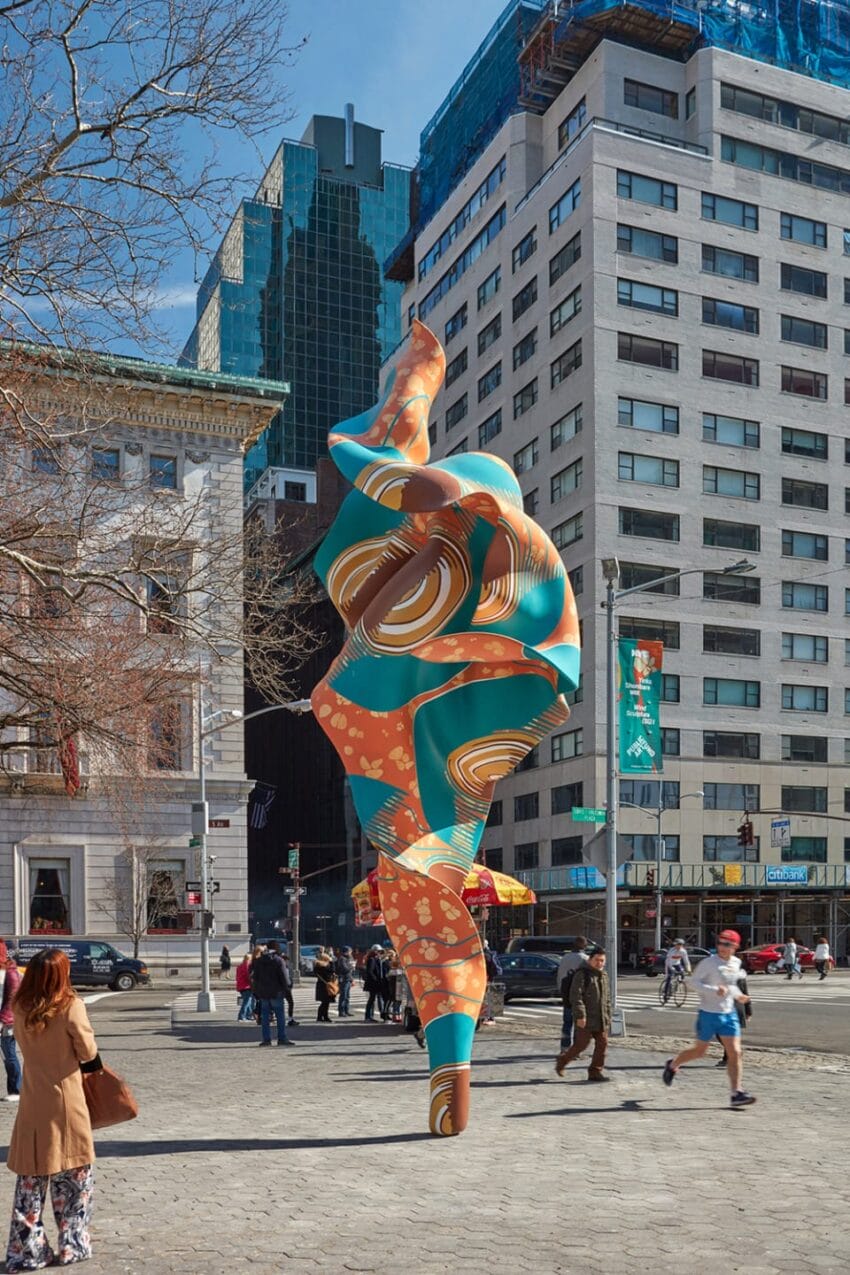 Colorful abstract sculpture stands prominently in urban plaza with people walking by and tall buildings in the background.