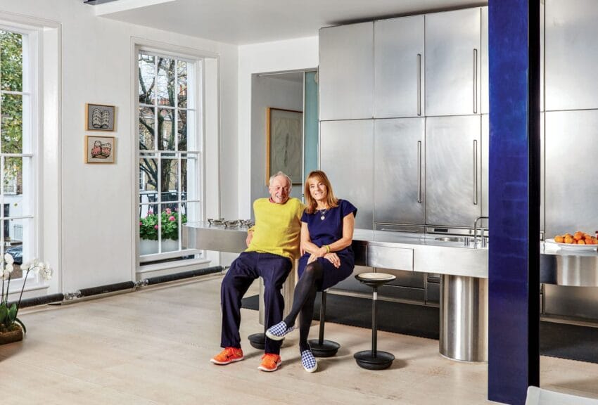 Two people sitting in a modern kitchen with stainless steel cabinets and large windows letting in natural light.