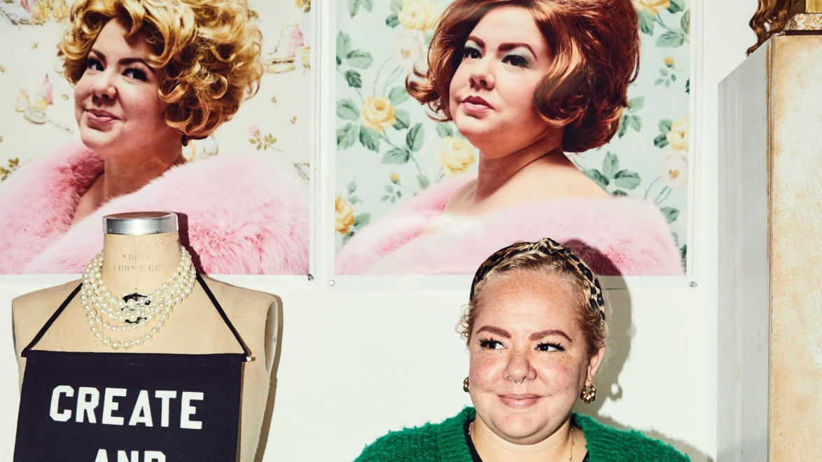 Person in green sweater beside mannequin with "CREATE AND RESIST" top, two vintage-style portraits on wall behind.