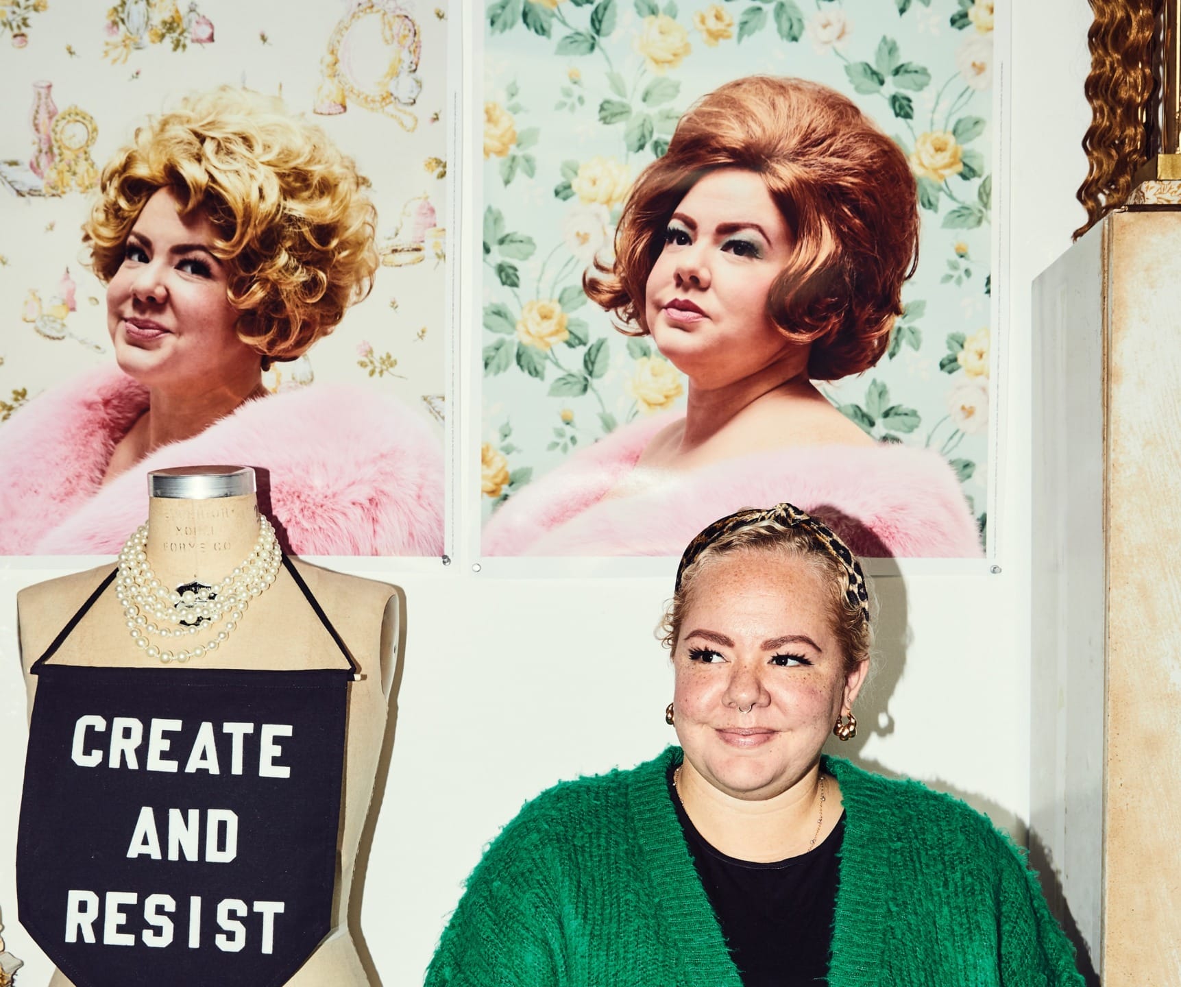 Person in green sweater beside mannequin with "CREATE AND RESIST" top, two vintage-style portraits on wall behind.