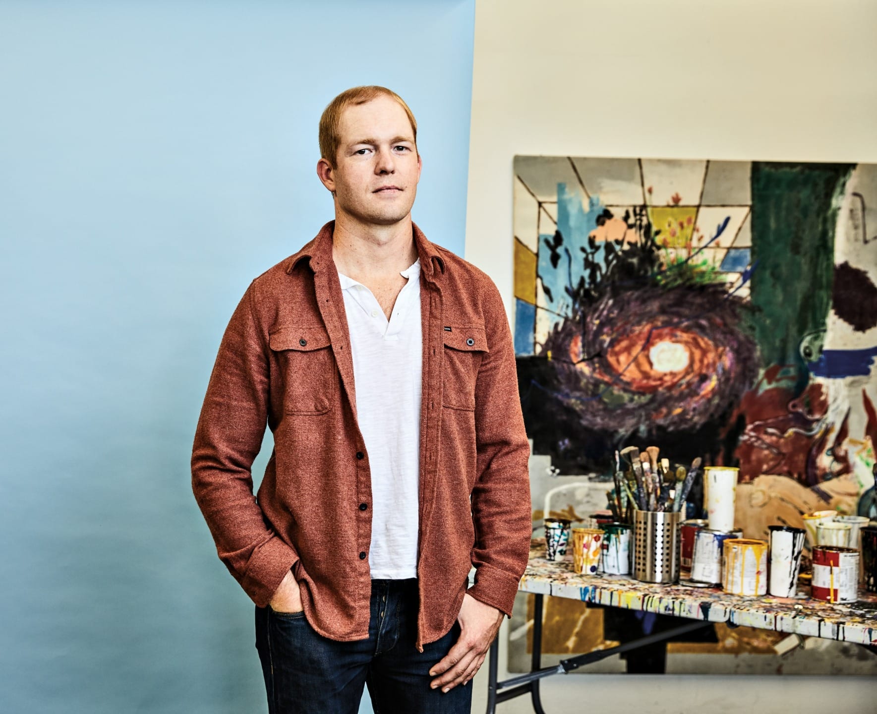 Man in a rust-colored jacket stands in an art studio with paintings and art supplies on the table behind him.