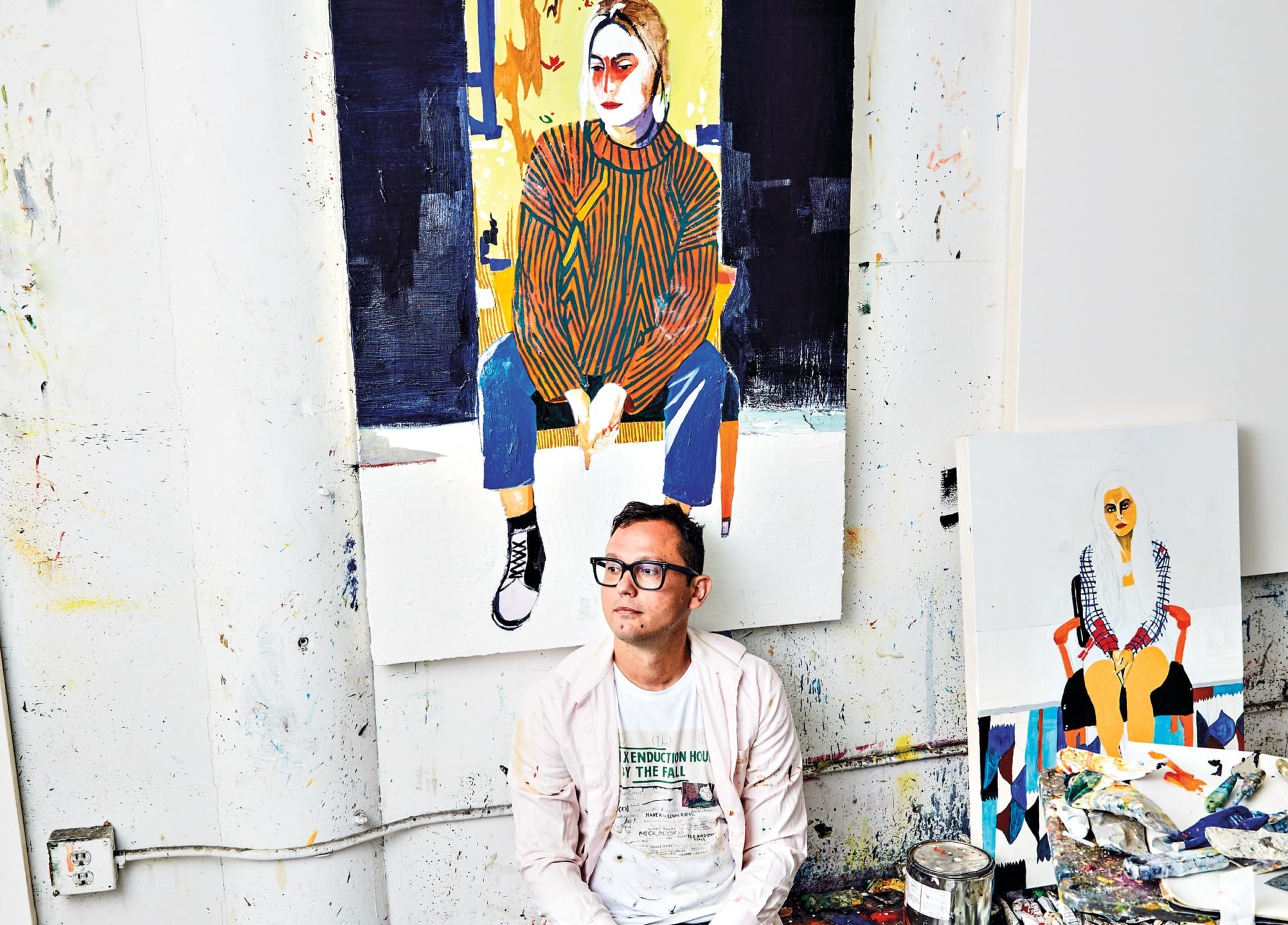 Artist sitting on floor in studio with colorful paintings of seated individuals hung on the wall.