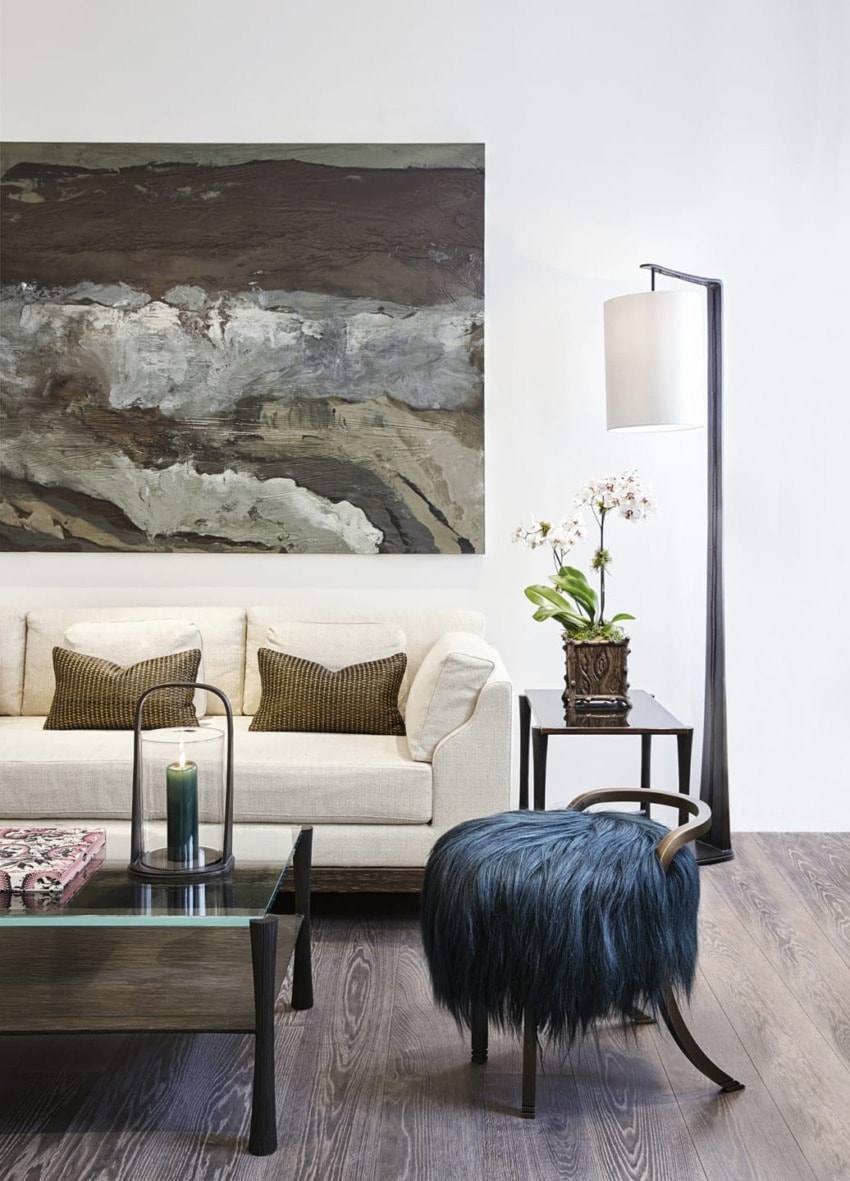 Modern living room with a beige sofa, abstract wall art, floor lamp, candle, plant, and a blue fur-covered stool.
