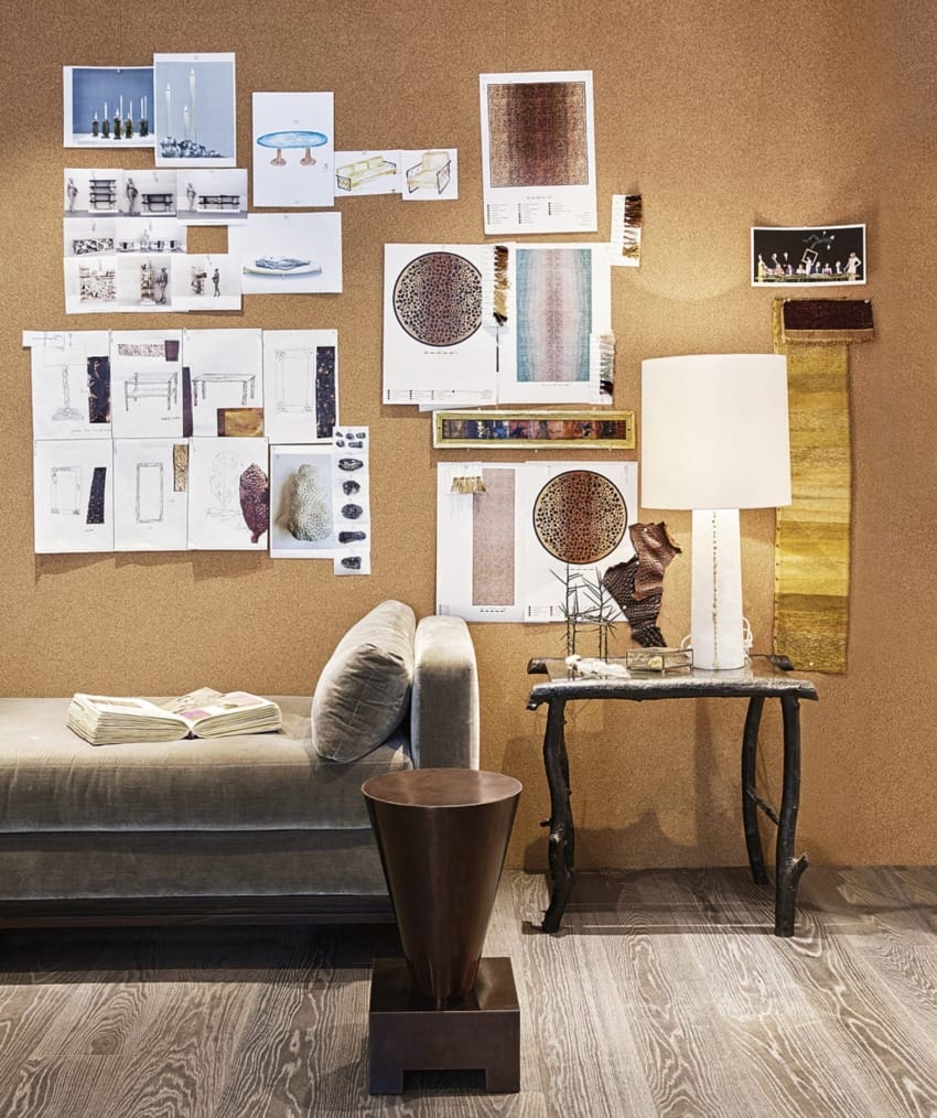 A cozy interior design studio with a couch, wall-mounted design plans, a unique table lamp, and a wooden stool on a textured floor.