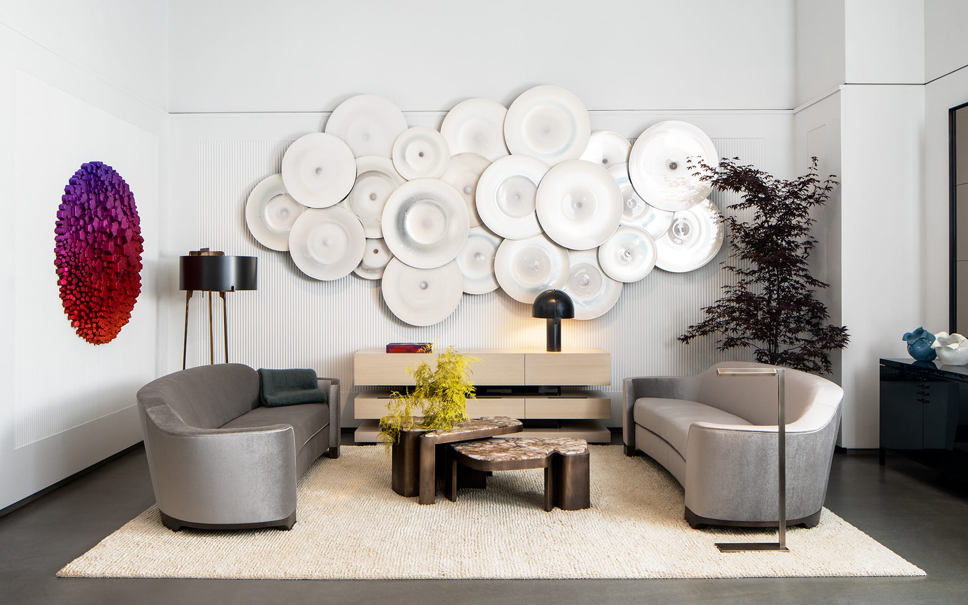 Modern living room with abstract wall art, gray sofas, circular coffee tables, black floor lamp, and decorative plants.