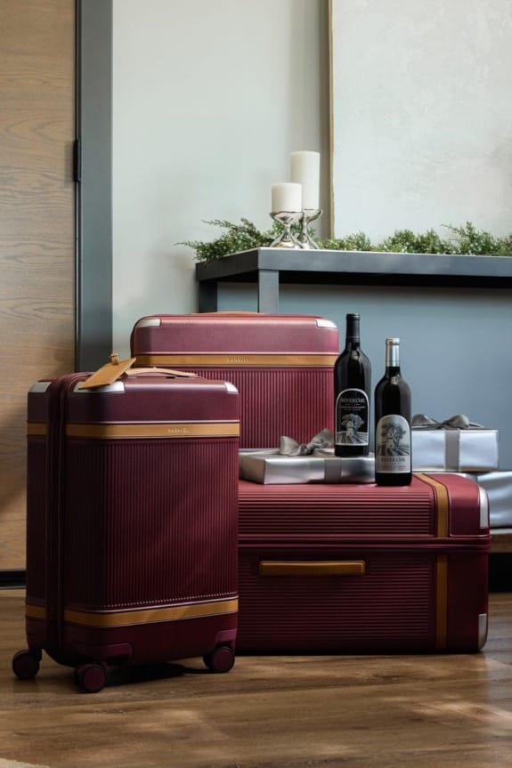 Three red suitcases with gold accents next to wine bottles and gift boxes in a stylish indoor setting.
