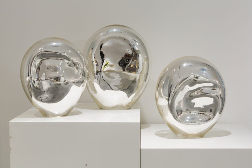 Three reflective silver glass sculptures on white pedestals against a plain background.