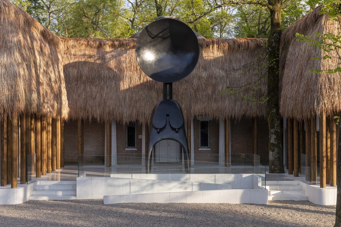 Large black sculpture with a horn shape stands in front of a thatched building surrounded by trees and glass barriers.