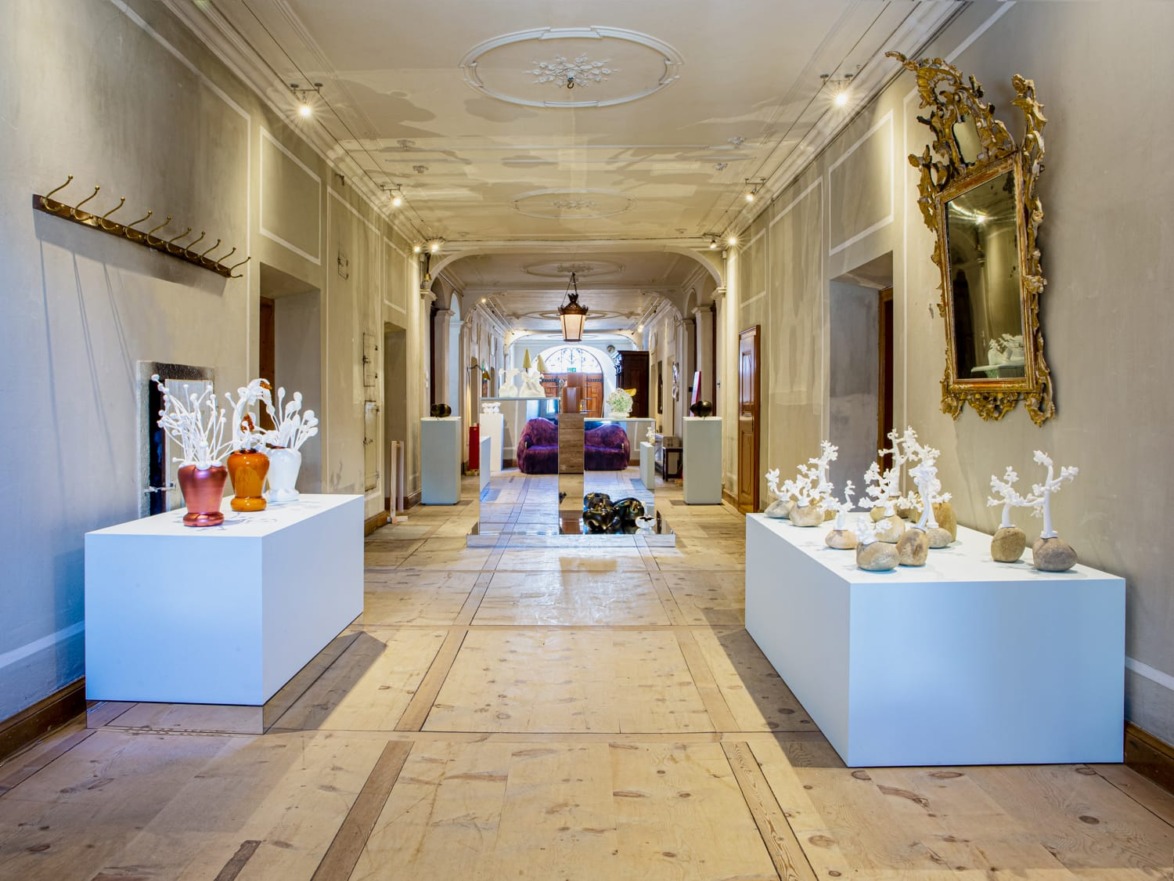 Art gallery hallway with modern sculptures on white pedestals, ornate mirror, and rustic wooden flooring.
