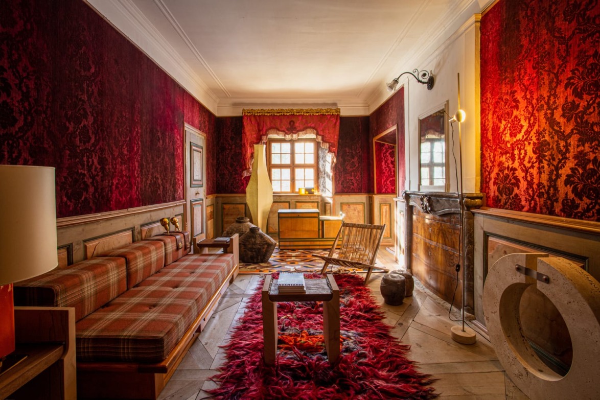 Vintage living room with red wallpaper, plaid sofa, wooden furniture, large window, and red rug on wooden floor.