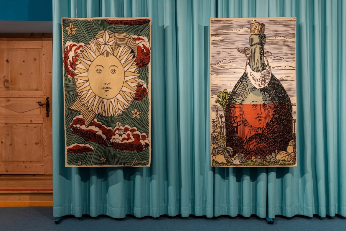 Two tapestries hanging on a teal curtain; one features a sun and clouds, the other a bottle with a red face inside.
