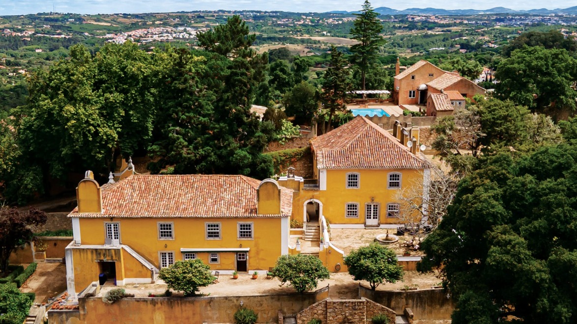 Aerial view of a historic yellow estate surrounded by lush green trees and rolling hills under a cloudy sky.