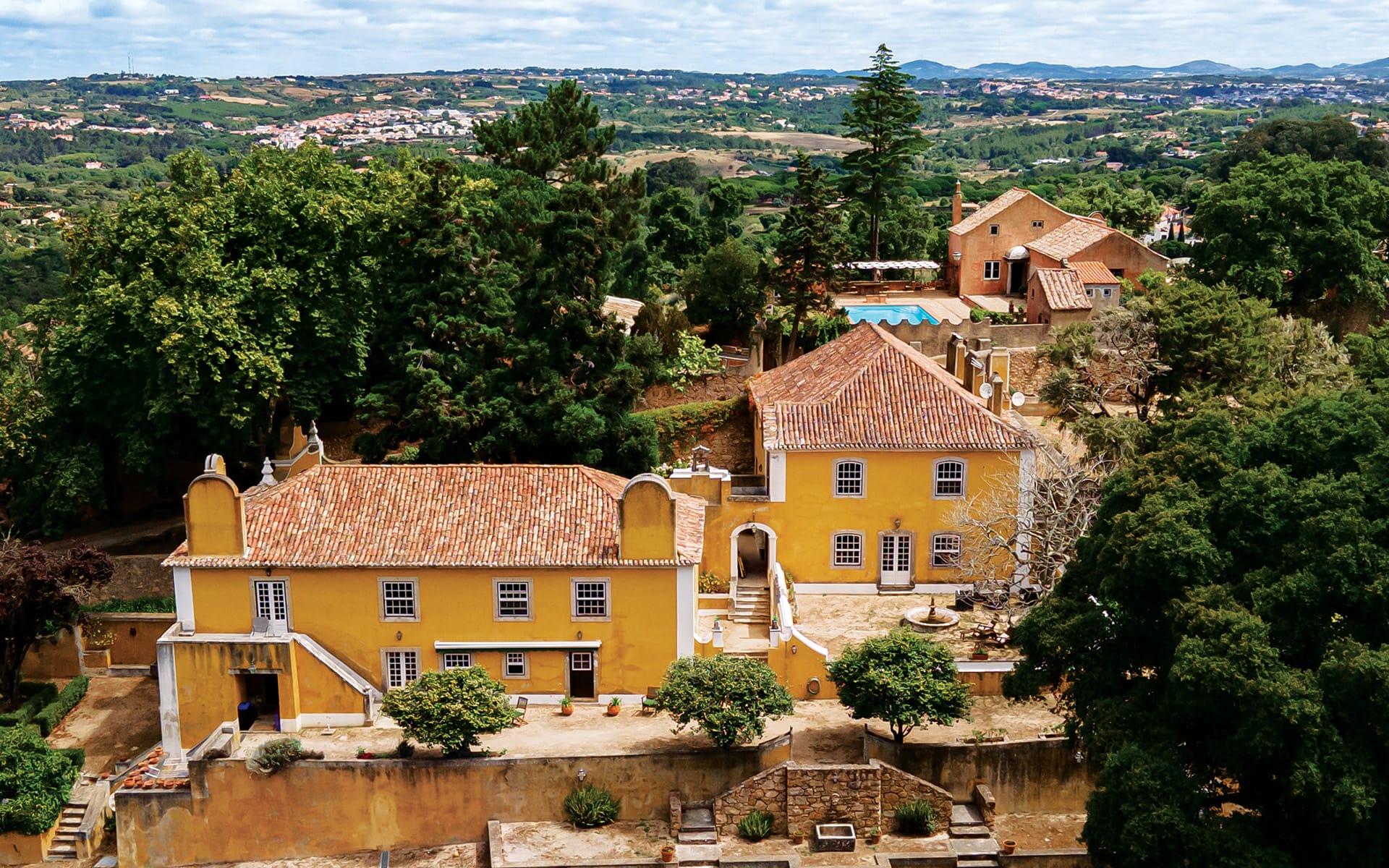 Aerial view of a historic yellow estate surrounded by lush green trees and rolling hills under a cloudy sky.