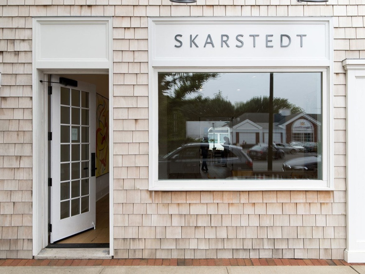 Front view of an art gallery with SKARSTEDT sign, featuring a large window and an open wooden door on a shingled exterior wall.