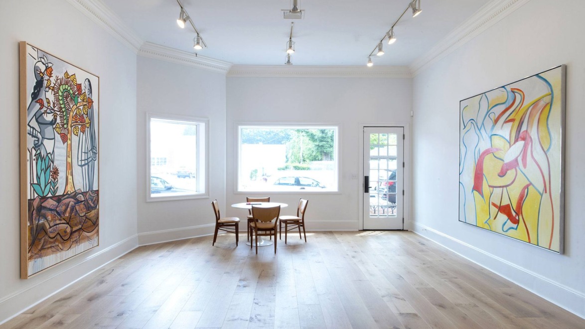Spacious art gallery with wooden floors, modern lighting, and colorful abstract paintings on white walls.