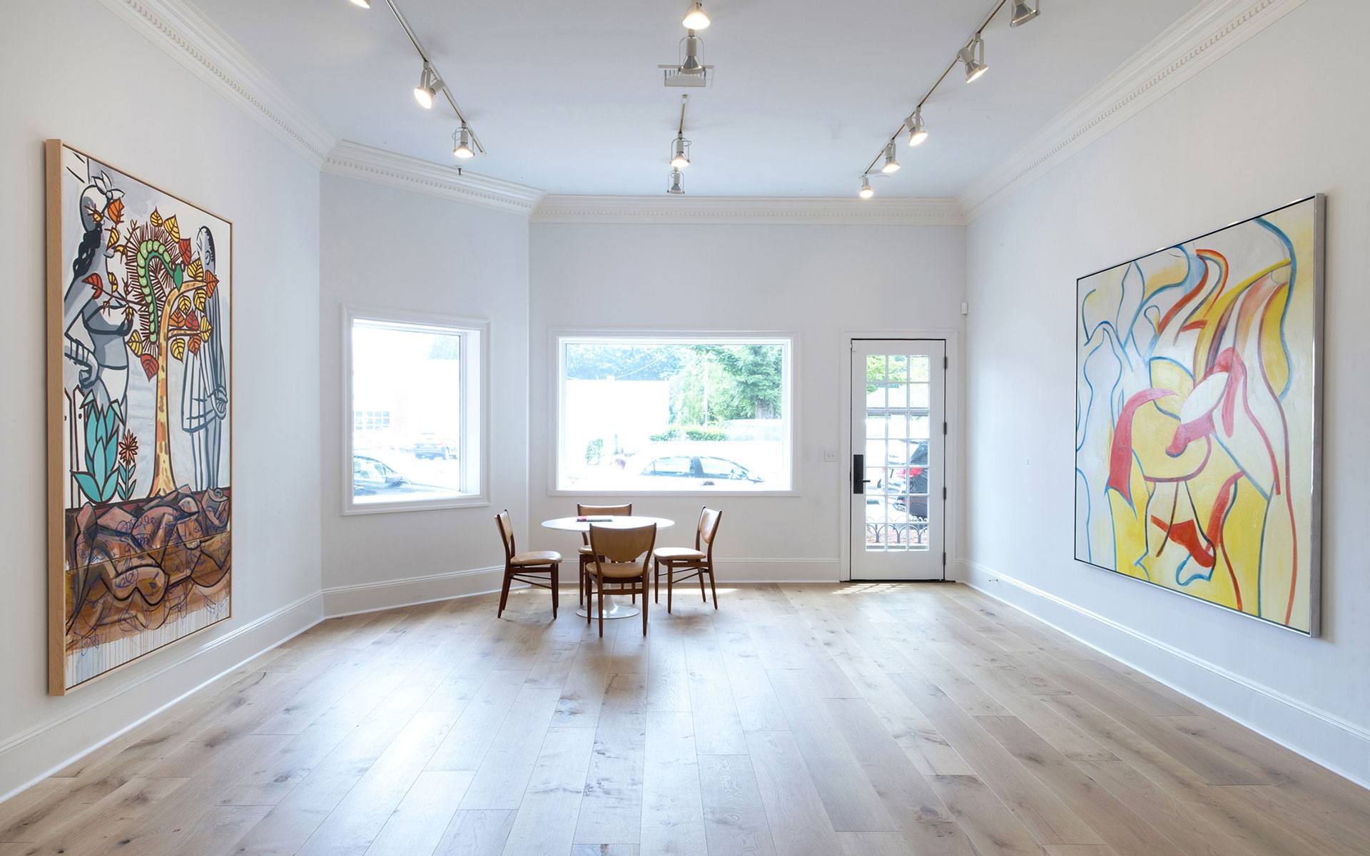 Spacious art gallery with wooden floors, modern lighting, and colorful abstract paintings on white walls.