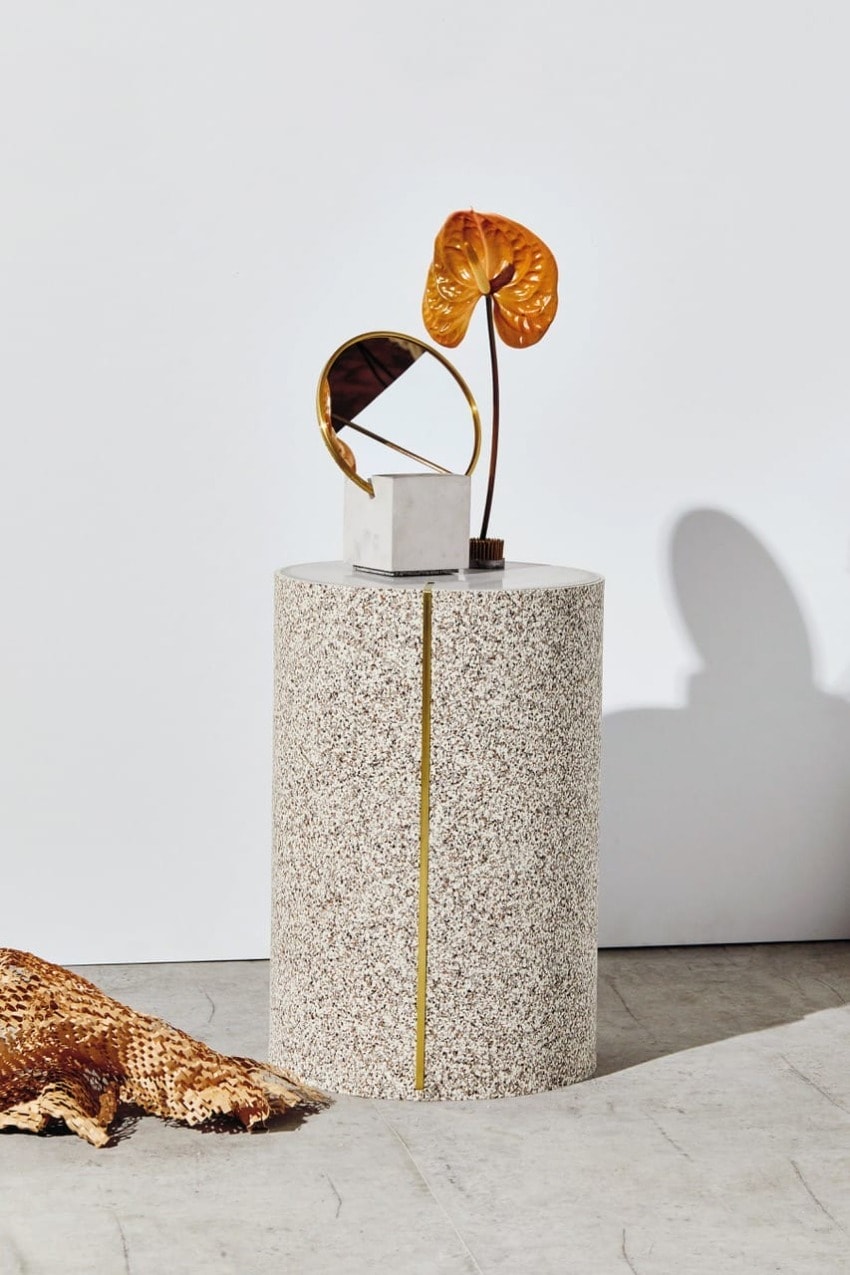 Decorative round speckled pedestal with gold accents, featuring a small mirror and an orange flower in a minimalist setting.