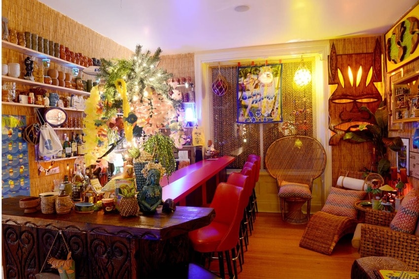 Cozy tiki bar with festive lights, tropical decor, bamboo accents, and a red counter lined with stools.