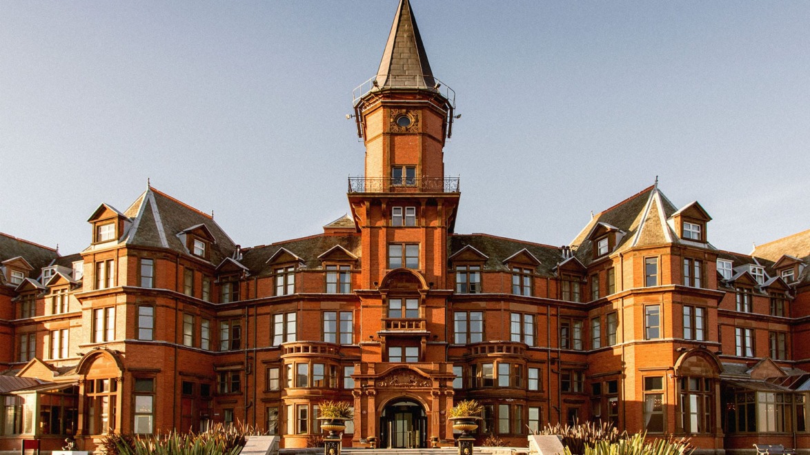 Historic red brick building with tall central tower and ornate architectural details against a clear blue sky.