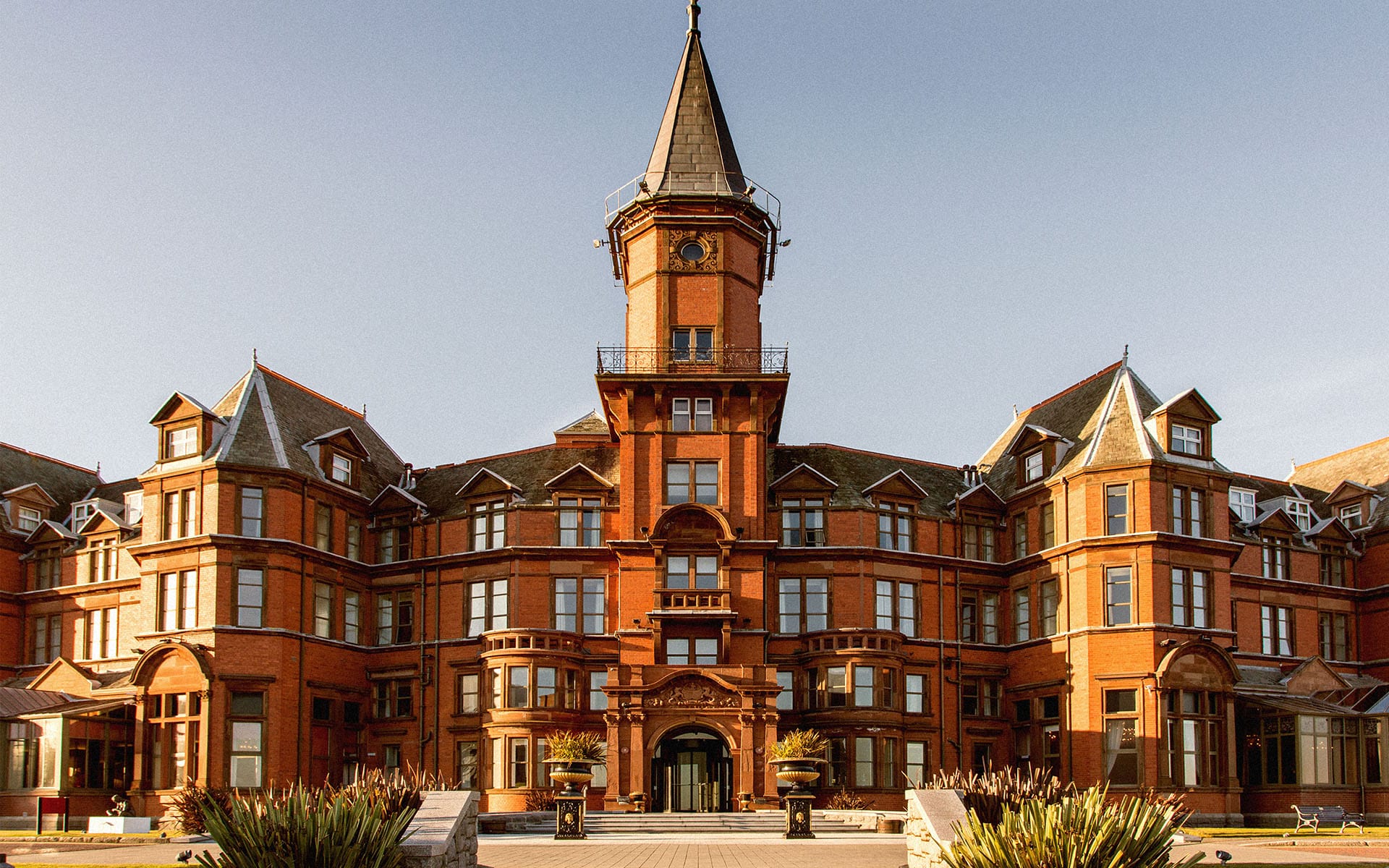 Historic red brick building with tall central tower and ornate architectural details against a clear blue sky.