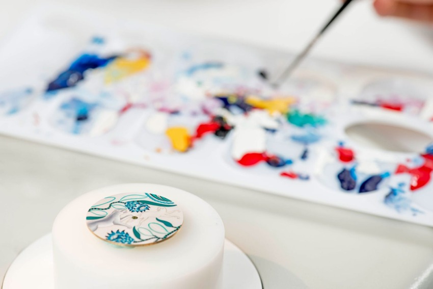 Cake decoration in progress with colorful paint palette and intricate floral design on a fondant disc.